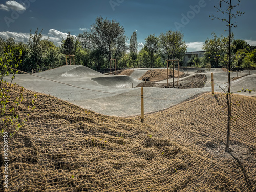 Sunny skatepark with concrete ramps and dirt hills, surrounded by trees and blue sky, vibrant and energetic scene