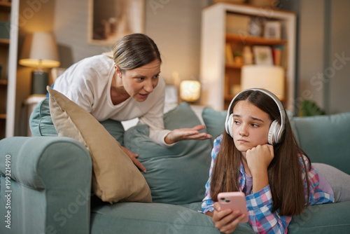Slika na platnu A young girl wearing headphones looks disengaged on a couch while a frustrated adult gestures and talks to her in a cozy living room setting