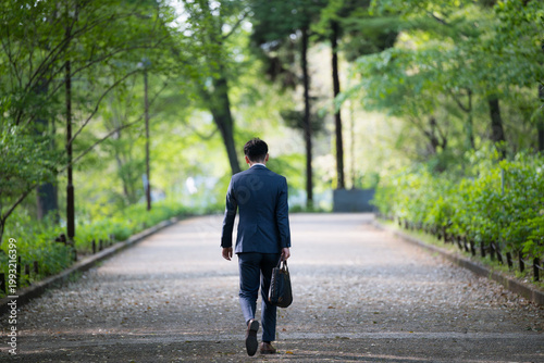 新緑の公園をビジネスバッグを持って歩く営業マンの仕事イメージ全身の広角　顔無しの後ろ姿、落ち込むネガティブストレスイメージ