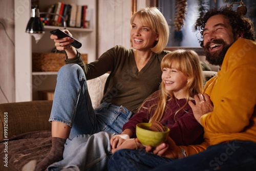 Three family members sit on a couch, laughing and watching a movie. The atmosphere is warm and cozy, filled with joy and relaxation as they share snacks.