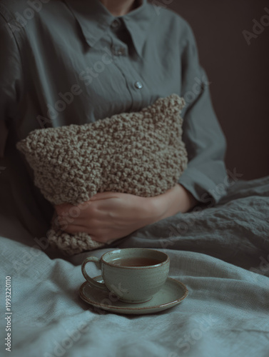 Herbal tea for cramps provides comfort and relaxation as person sits in bed holding knitted pillow, creating soothing and calming atmosphere for relief and wellness