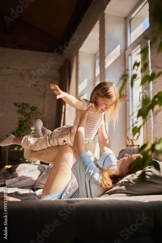 фотографія Mother playing airplane game lifting daughter on legs in sunny living room