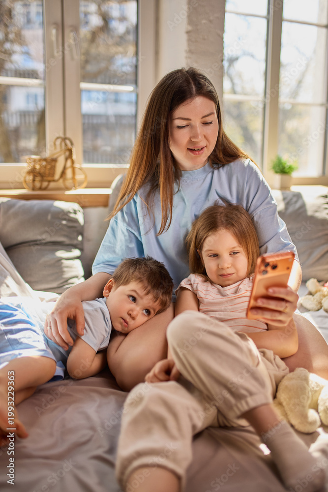 Obraz premium Mother and children using smartphone together on sofa in sunlit living room. Concept of mobile app marketing, digital communication and online content consumption in family environment.