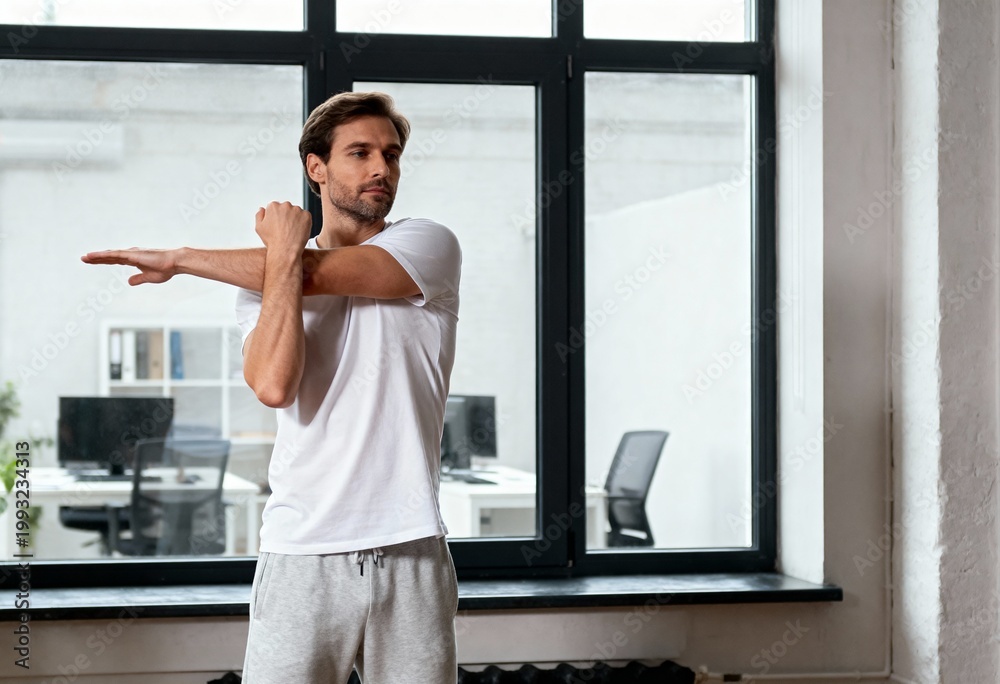 Obraz premium Focused man in sportswear stretching his arm in a modern office. Workplace wellness and active break concept with copy space