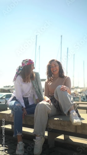 Two young women laughing and talking while sitting at a sunny marina with boats in the background