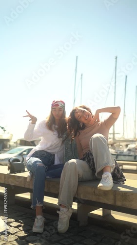 Two cheerful young women having fun together sitting on a bench at the city marina on a sunny day