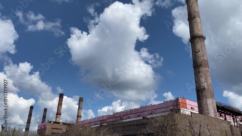 Old industrial factory with tall chimneys against blue sky with clouds. Camera moves upward from low angle. Concept of industry, pollution, energy production and urban infrastructure. 4K video.