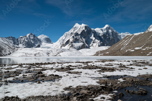 A serene winter landscape of Gurudongmar, North Sikkim, featuring snow-dusted stones in the foreground against a backdrop of majestic, snow-capped Himalayan peaks during a snowfall.