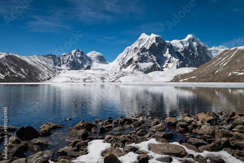 A crystal-clear view of Gurudongmar Lake in North Sikkim, with submerged stones visible through the transparent turquoise water in the foreground.
