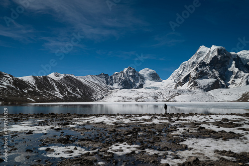 A solitary man stands in the vast, desolate landscape of Gurudongmar, North Sikkim, gazing toward snow-capped Himalayan peaks under a dramatic sky.