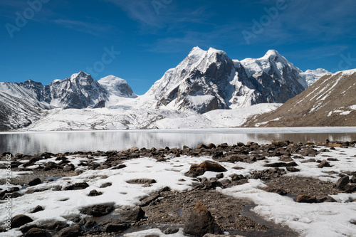 A serene winter landscape of Gurudongmar, North Sikkim, featuring snow-dusted stones in the foreground against a backdrop of majestic, snow-capped Himalayan peaks during a snowfall.