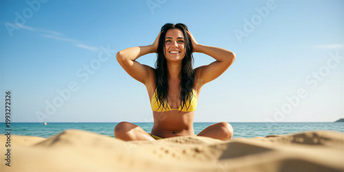 Slim brunette sitting on sandy beach in yellow bikini, laughing and showing perfect teeth, hands in hair, with bright blue ocean and clear blue sky in the background.