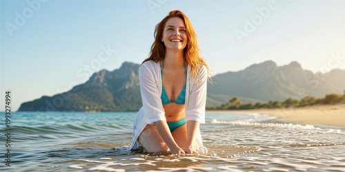 Slender red-haired woman wearing a blue bikini and white shirt, kneeling in the shallow water of a beach, with mountains visible on the horizon behind her.