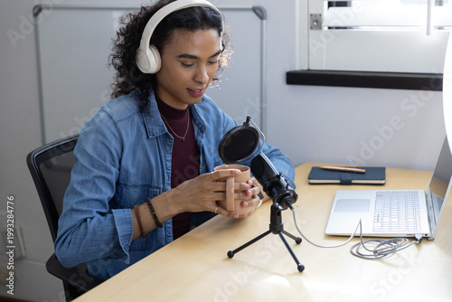 Nonbinary person in twenties in denim at desk holding mug, recording with microphone and headphones
