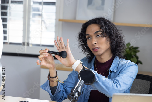 Non-binary creator wearing denim jacket showing makeup brush and speaking toward mic at home studio
