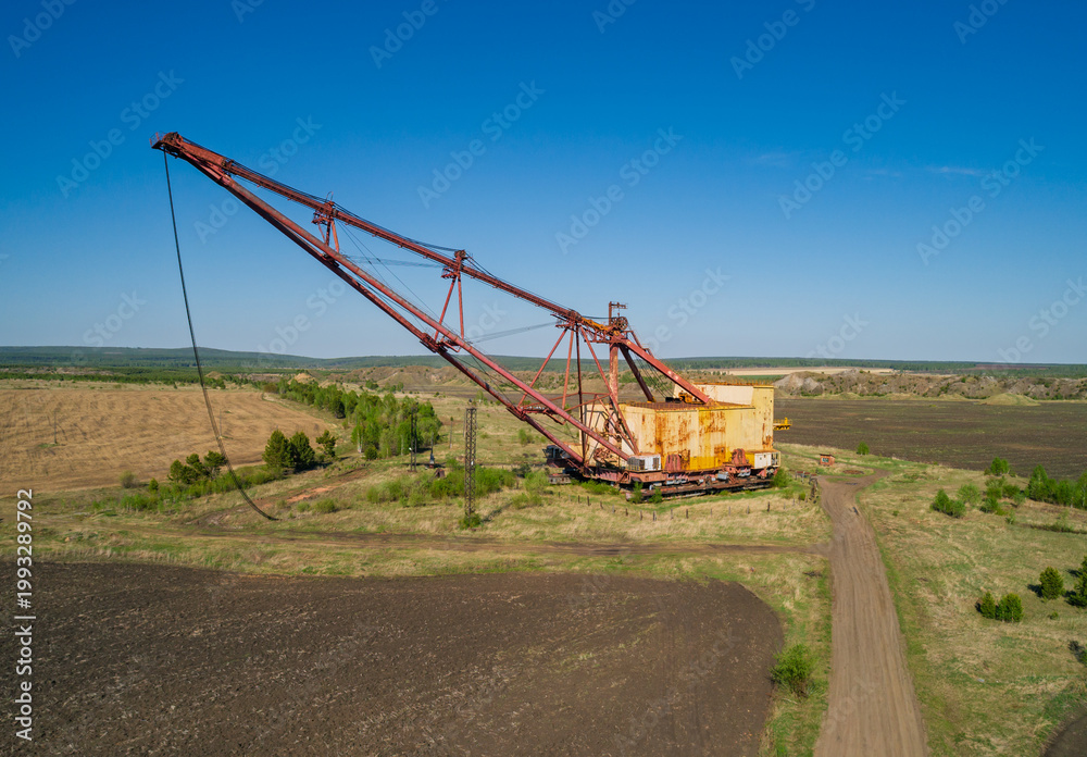 Fototapeta premium Massive industrial dragline excavator rests in an open field, surrounded by sparse vegetation and dirt roads