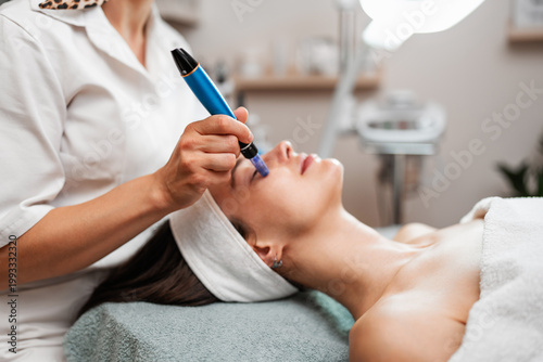 A professional cosmetologist uses a dermapen for a microneedling facial procedure on a young woman lying down in a beauty clinic.