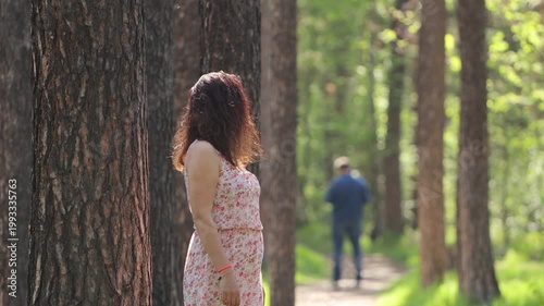 A woman and a man in a forest. A young woman stands leaning against a pine tree trunk. Behind her on the path is the silhouette of a man. She looks back and stares at him for a moment.