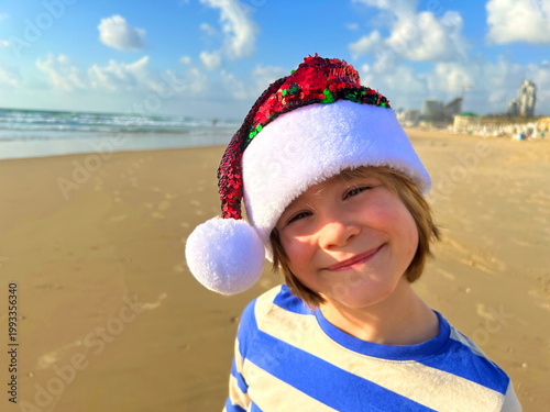 Christmas on the island! A boy in a Santa Claus hat on the beach by the sea. Winter holidays, family vacation