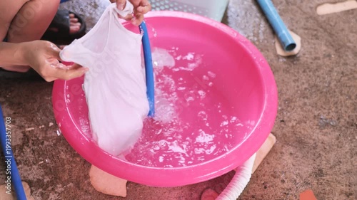 Close-up of hands rinsing and squeezing a white cloth in a pink water basin at home. The image reflects hand washing, laundry care, household chores, hygiene, cleaning, and everyday domestic routine.