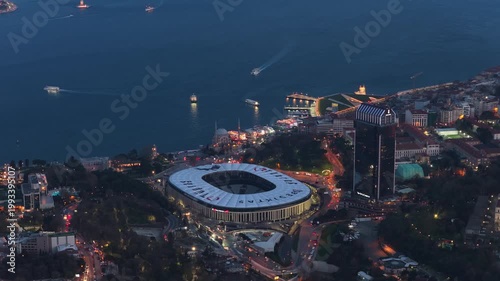 High angle scene of Istanbul at twilight, showing illuminated Vodafone Park stadium, dark waters of Bosphorus Strait with boats, and bustling city streets