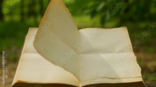 The video depicts an open book placed outdoors, likely on a flat surface such as a table or the ground, with a natural, green, and slightly blurry background suggesting a garden or park setting