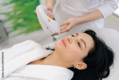 Tableau sur toile Middle-aged woman receiving relaxing rf lifting procedure for face and neck in beauty salon, cosmetologist using applying professional equipment for non invasive facial treatment