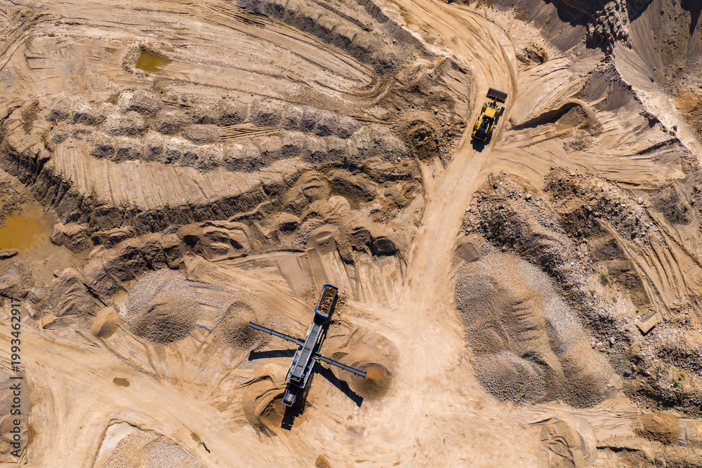 Fototapeta premium Aerial view of an open pit quarry shows a yellow haul truck climbing a dirt ramp. A mobile crusher with conveyors sits central amid gravel and sand stockpiles.
