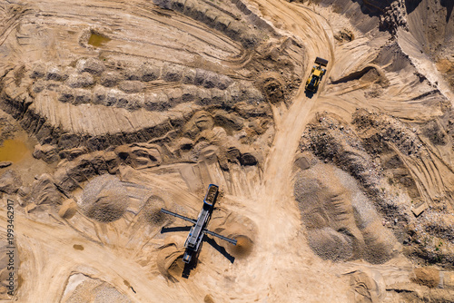 Aerial view of an open pit quarry shows a yellow haul truck climbing a dirt ramp. A mobile crusher with conveyors sits central amid gravel and sand stockpiles.