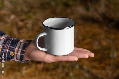 Mock-up of a white enamel mug with a rim on a hand in a flannel shirt, a metal cup for hot drinks, coffee, tea.