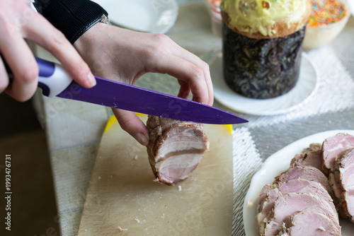 A woman is cutting ham on the table. Easter eggs are on the background.