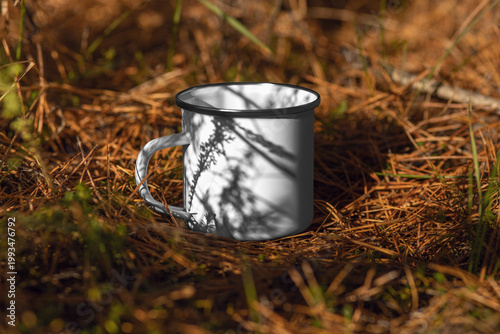 Template of enameled white mug with iron rim, handle, with shadows from branches, on a background of pine needles, outdoors.