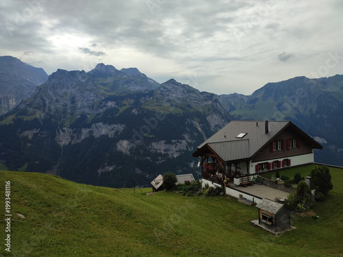 Charming countryside house on a green hillside overlooking majestic alpine mountains. Scenic landscape with cloudy sky, lush grass, and peaceful rural atmosphere.