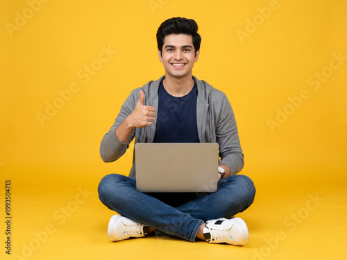 young indian male student sitting cross legged with laptop showing thumbs up isolated on yellow background representing online learning education success and college admission concept