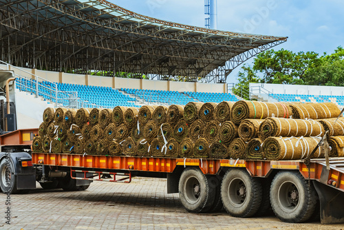 Truck loaded with sod rolls parked at stadium for lawn landscaping and renovation for football field. Large shipment of natural sod rolls on heavy duty truck trailer with stadium bleacher background.
