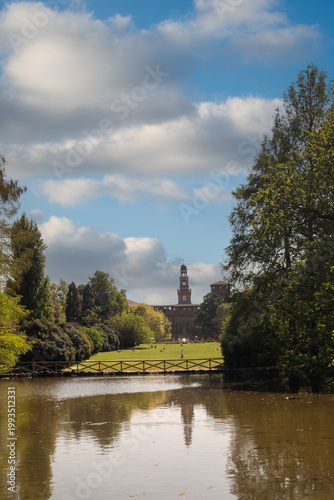 ​A scenic view of Parco Sempione in Milan featuring a peaceful pond with reflections, a wooden bridge, and the distant tower of Castello Sforzesco under a cloudy sky.
