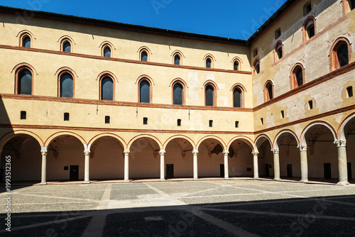 The historic courtyard of Castello Sforzesco in Milan features impressive Renaissance arcades with elegant columns and arched windows set against a vibrant blue sky