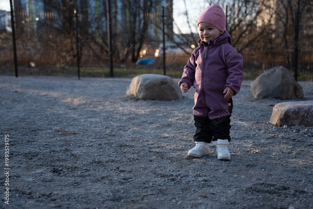Fototapeta premium smiling toddler in a purple snowsuit and pink beanie stands on gravel at golden-hour in a quiet urban park playground, soft backlight, scattered boulders and bare trees evoke calm, chilly atmosphere
