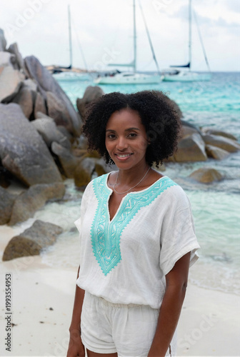 A smiling Black woman in a white and turquoise top stands on a tropical beach.