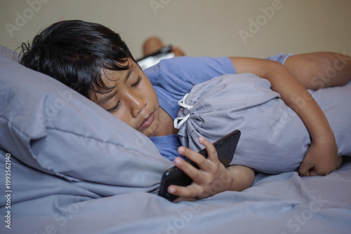 Foto Young boy lying in bed hugging a bolster while using smartphone with selective focus