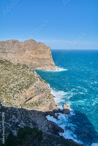 The steep coastal landscape of Cap de Formentor features jagged rock formations and white waves crashing against the base of the cliffs.