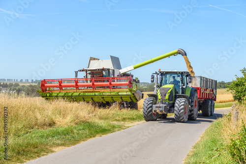 Combine harvester loading a trailer pulled by a tractor with barley grains on a clear summer day