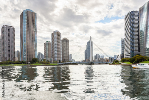 High rise apartment buildings on the banks of a river under cloudy sky in autumn. A modern suspension bridge connecting the two sides of the river is visible in distance. Tokyo, Japan.