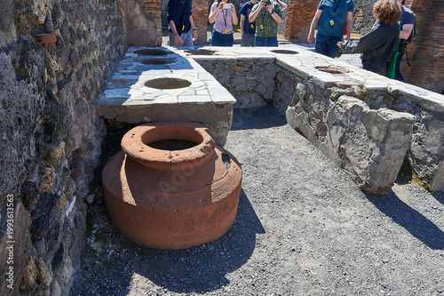 “Pompeii: Ancient thermopolium counter with amphorae and tourists in Roman street food setting”