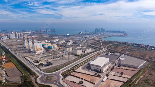 Aerial view of large coastal industrial zone featuring combined cycle gas turbine power plant with striped exhaust stacks, high-voltage substation, water desalination station, concrete plant, LNG term