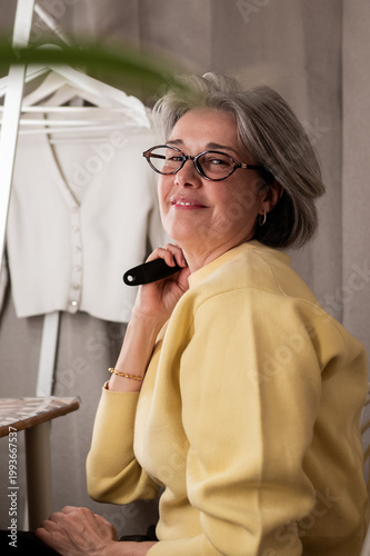 Mature woman with gray hair wearing glasses and a yellow sweater, holding a tailor's tool, looking at the viewer and smiling