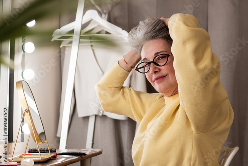 Mature woman with eyeglasses and a bright yellow sweater thoughtfully styling her grey hair, preparing for the day at a dressing table illuminated by lights
