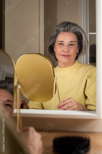Senior woman with silver hair smiling into a golden vanity mirror, practicing self-care and confidence while reflecting on aging and morning routine in her cozy home