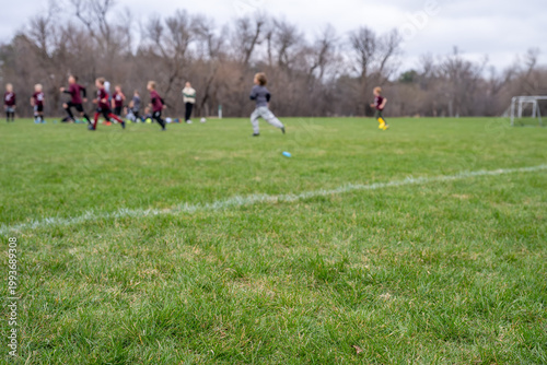 Kids play soccer on a grassy field during a cloudy day in a local park with other players in the background