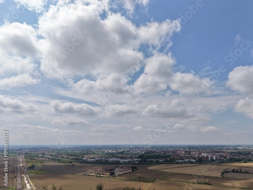 Obraz na plátně Cloudscape and Countryside: An aerial vista of a rural landscape, where puffy white clouds drift gracefully across a radiant blue sky, overseeing the patchwork of fields and the gentle horizon